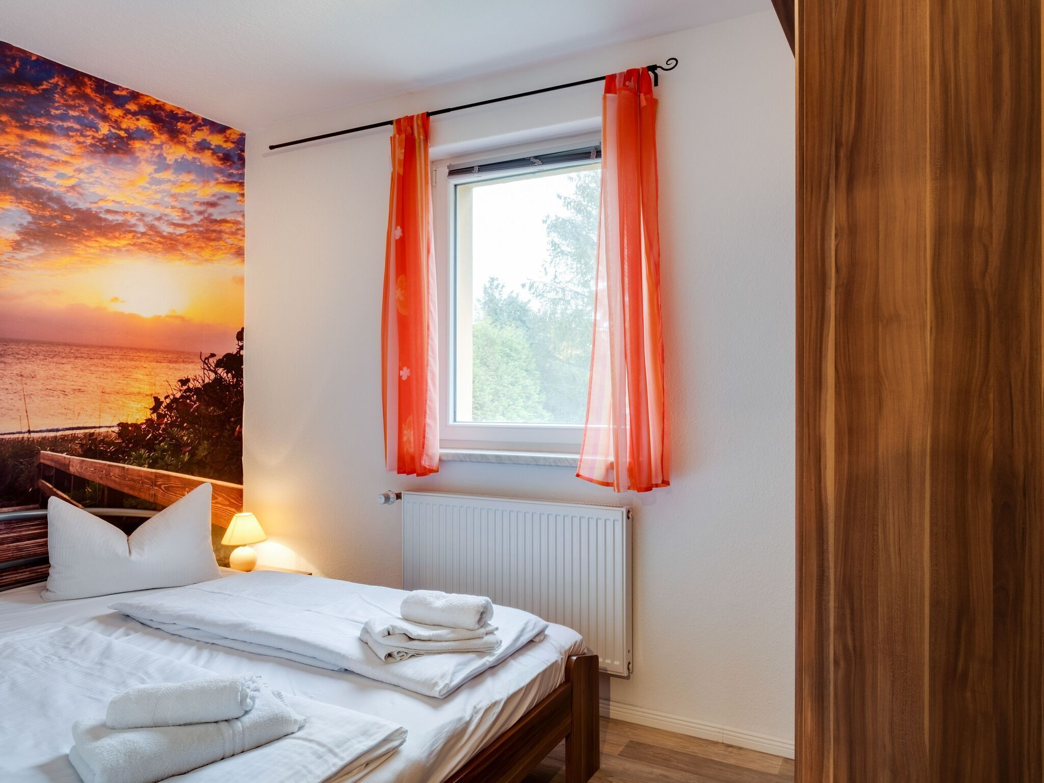 A bedroom with a bed, white bedding, a window with orange curtains, a radiator, and a sunset mural on the wall.