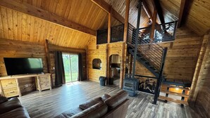 Interior de una cabaña de troncos con piso de madera, un sofá de cuero, un televisor y una escalera.