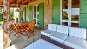 Un patio exterior con mesa y sillas de madera, un sofá blanco y contraventanas verdes en las paredes.