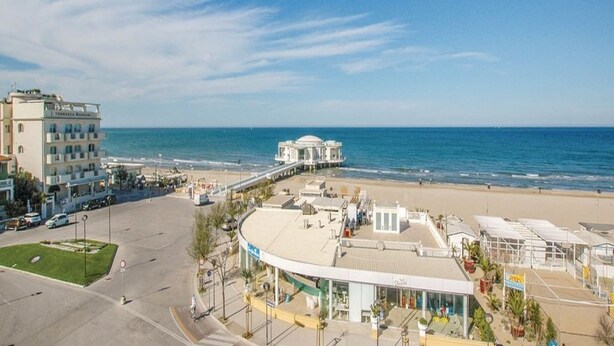 Un hotel frente al mar con vista a la playa, zona de estacionamiento y un pequeño paseo marítimo.