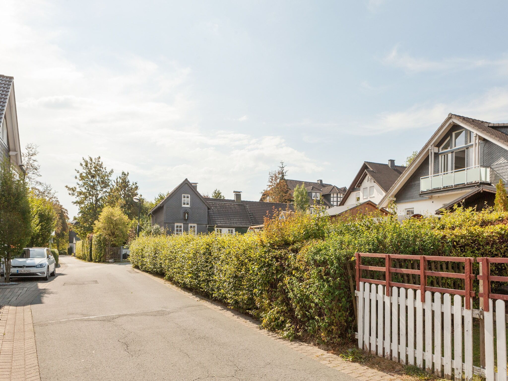 A residential street with houses, a white car parked on the side, and a hedge along the road.