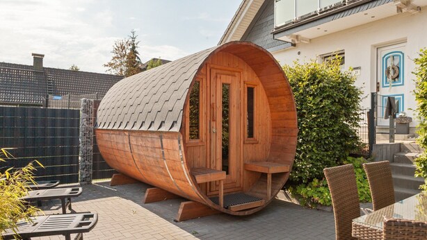 A wooden barrel-shaped structure with a door and windows, situated in an outdoor patio area with wicker furniture and a paved floor.
