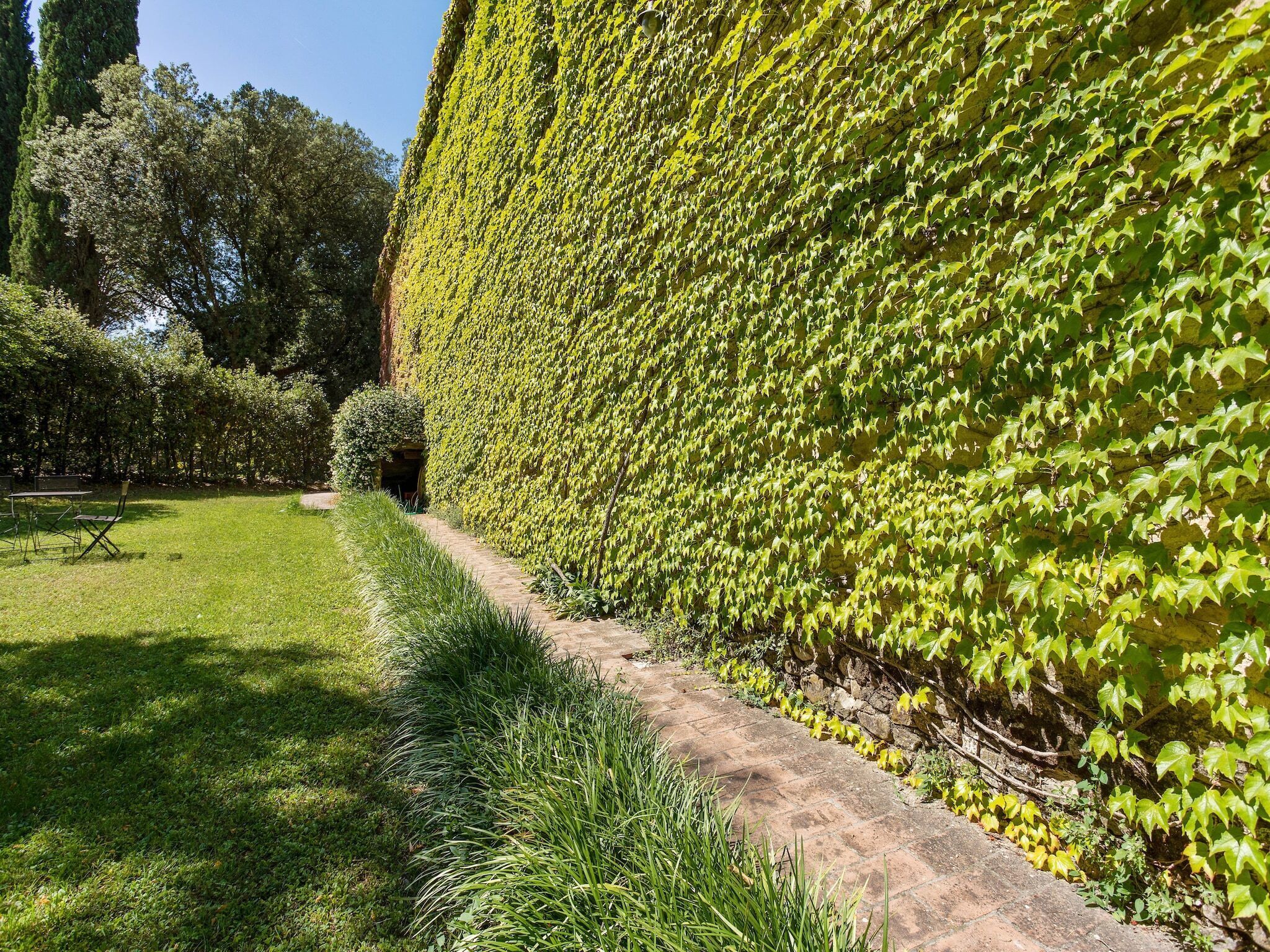 Un jardín con un sendero de piedra, un seto cuidado y un pequeño juego de mesa y sillas.