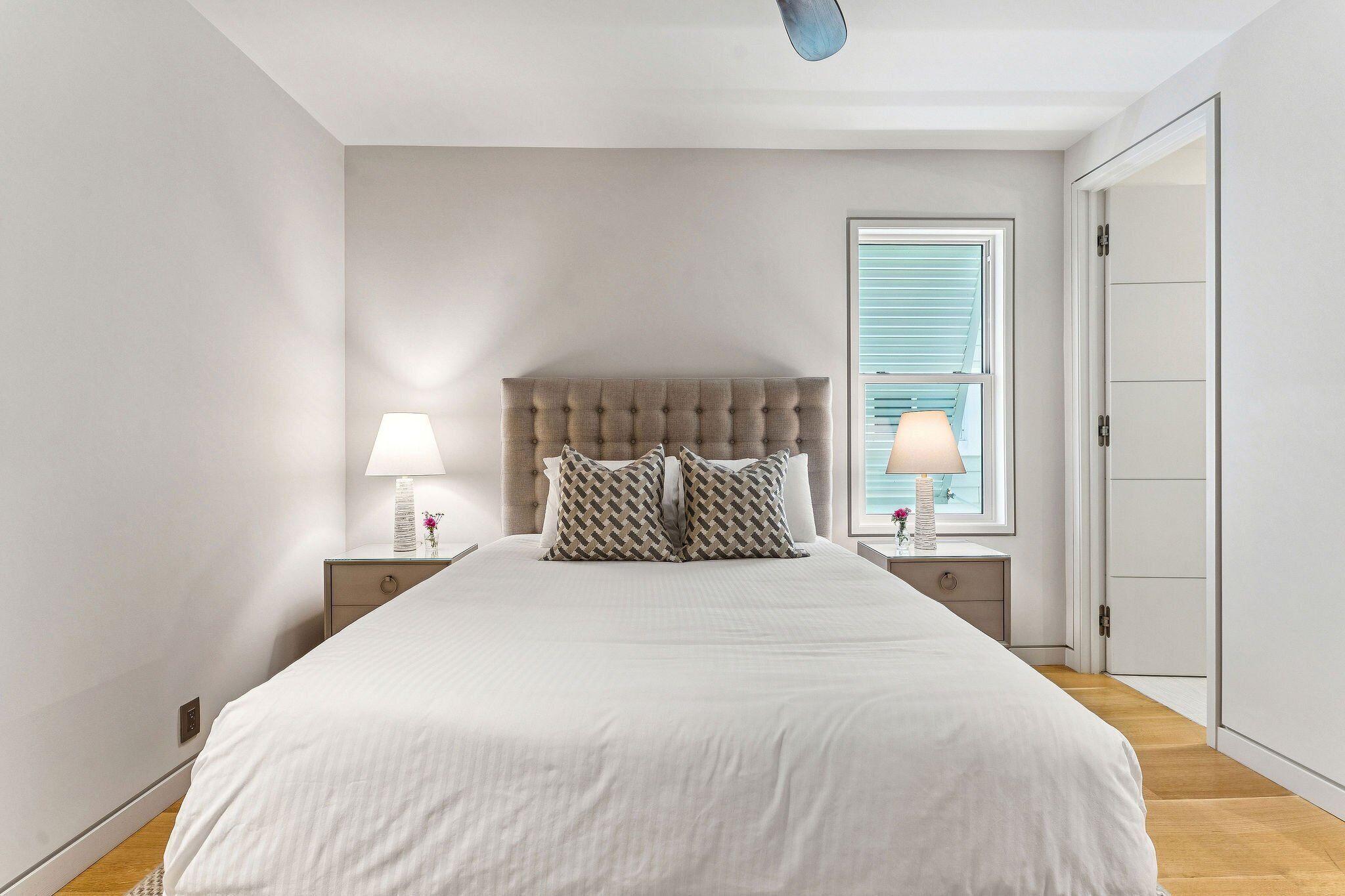 A bedroom with a large bed, two bedside tables with lamps, a window with blinds, and a door.
