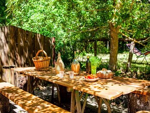 Une table de pique-nique en bois installée à l’extérieur, avec un panier, des bouteilles et un saladier de tomates.