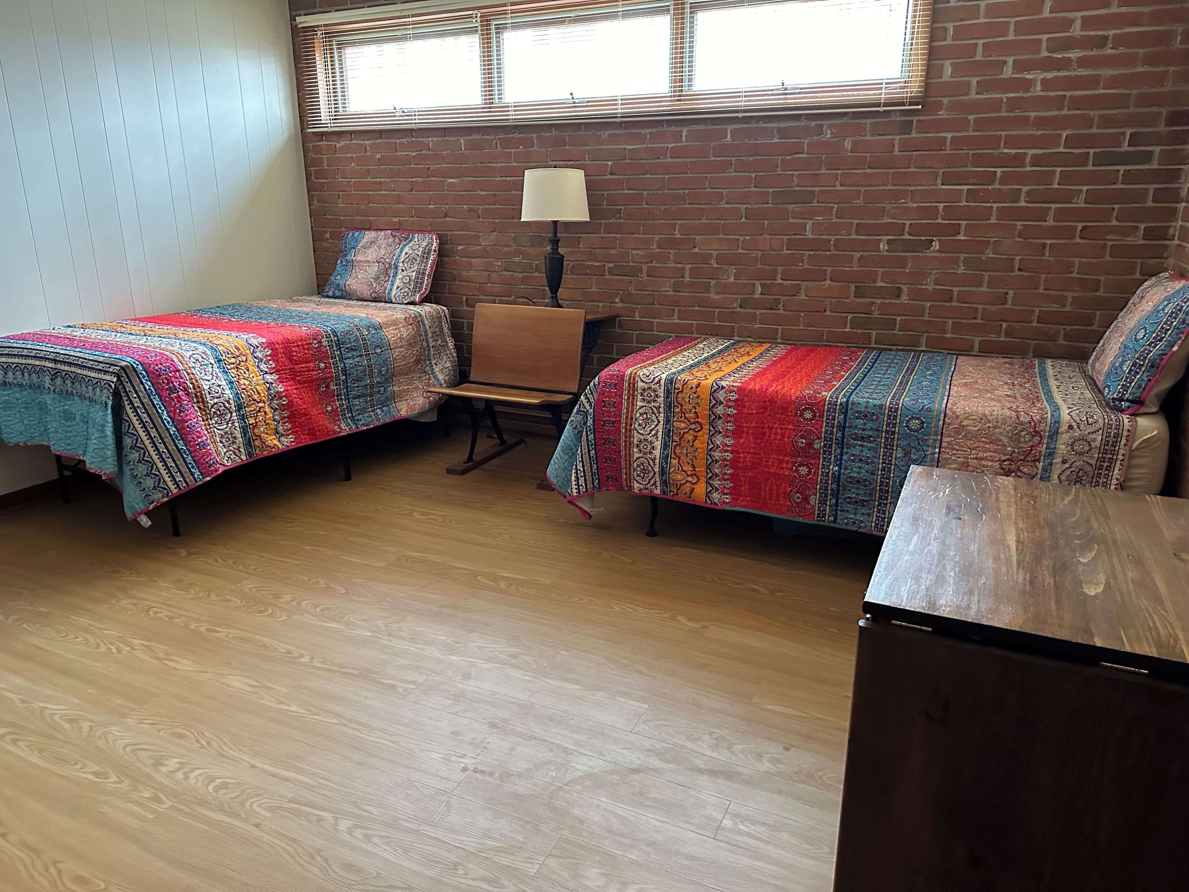 Una habitación con dos camas, una mesita, una lámpara y un muro de ladrillo.