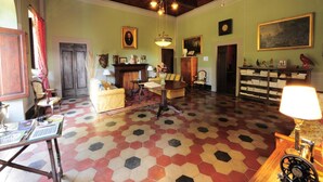 A spacious room with a hexagonal patterned floor, wooden ceiling beams, a chandelier, a fireplace, and a bookshelf filled with books.