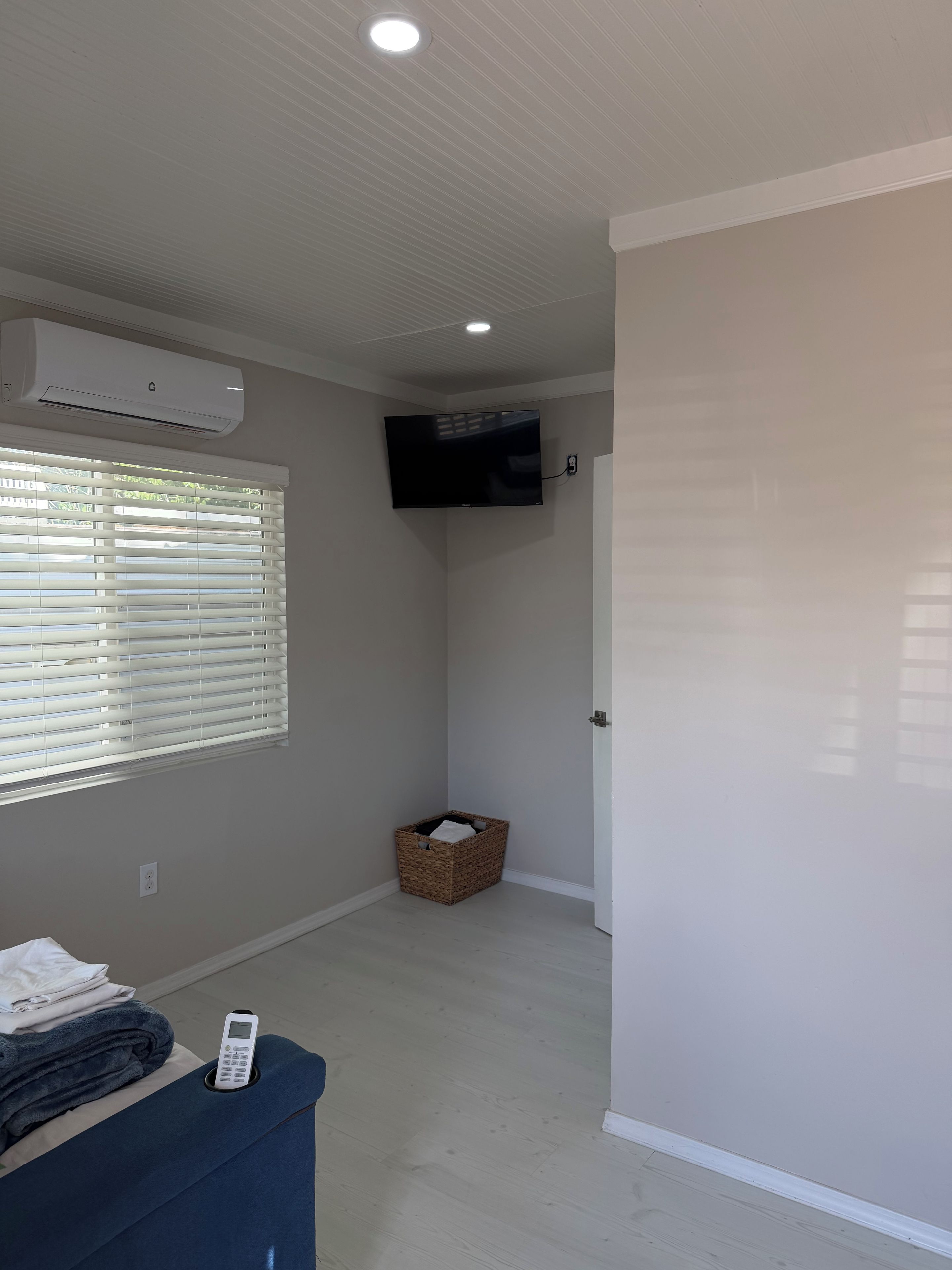 Una habitación con una televisión de pantalla plana montada en la pared, un calentador portátil, una cesta y una cama con un control remoto.