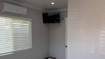 Una habitación con una televisión de pantalla plana montada en la pared, un calentador portátil, una cesta y una cama con un control remoto.