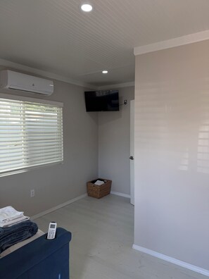 A room with a flat-screen TV mounted on the wall, a standing heater, a basket, and a bed with a remote control on it. - Tiny Home by The River (Los Lunas)