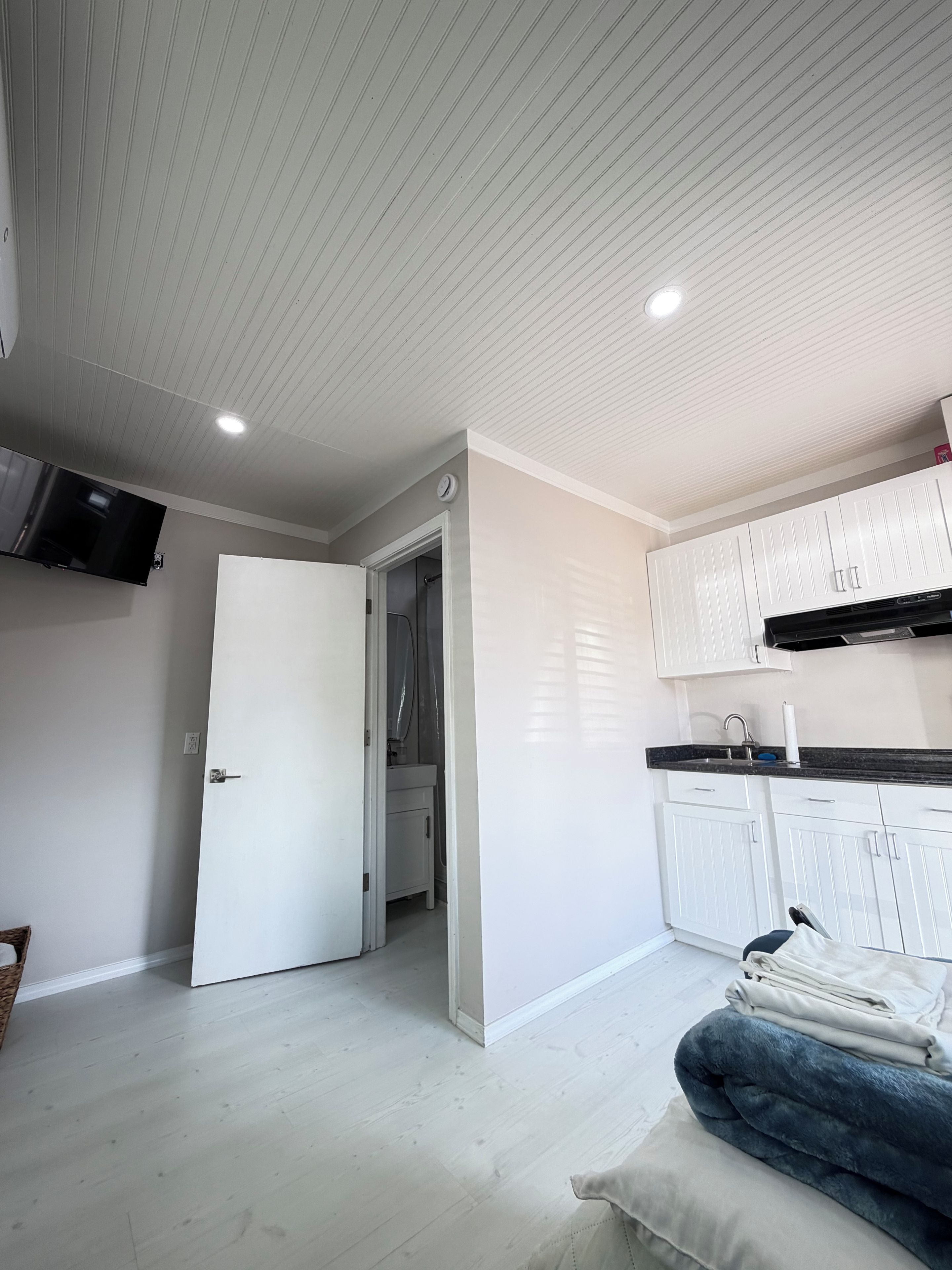 Una cocina moderna con armarios blancos y encimera negra, un televisor de pantalla plana sobre la encimera y una puerta que da a un baño.