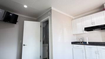 Una cocina moderna con armarios blancos y encimera negra, un televisor de pantalla plana sobre la encimera y una puerta que da a un baño.