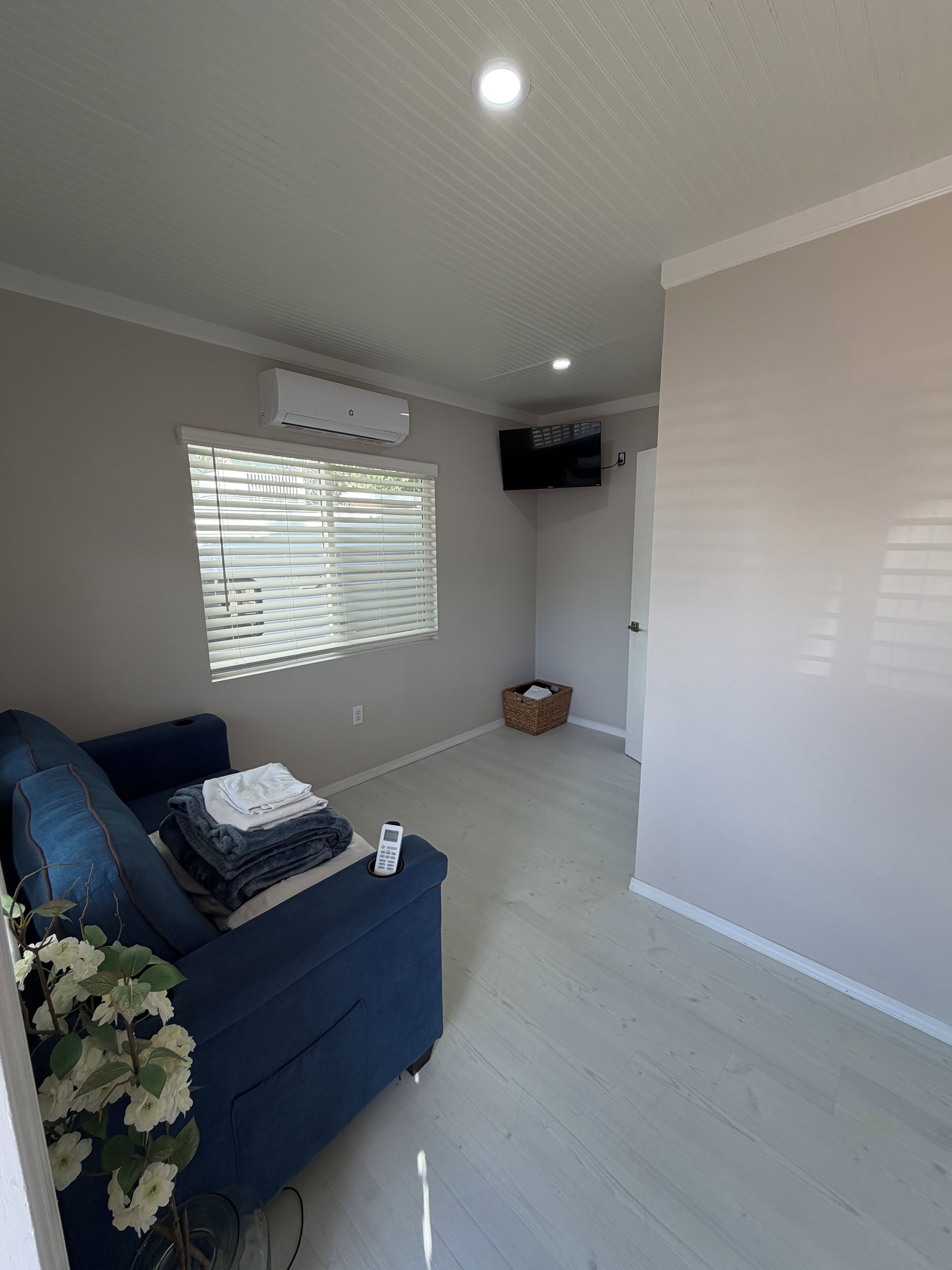 A living room with a blue sofa, a flat-screen TV mounted on the wall, and a window with blinds.