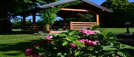 A garden with a wooden gazebo, a potted plant with pink flowers, and a grassy area.