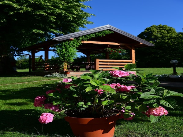A garden with a wooden gazebo, a potted plant with pink flowers, and a grassy area. - Apartment near Plitvička Lake and Korana River (Selište Drežnićko)
