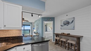 A kitchen with a black countertop, white cabinets, and a stainless steel sink. There is a wooden table with two stools and a painting of a turtle on the wall. - Sundunes 133 in Navarre Beach | Beachfront | Pool | My Beach Getaways (Navarre)