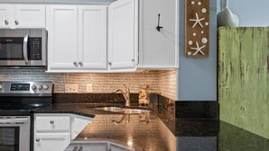 A kitchen with white cabinets, a stainless steel microwave, a double sink, and a backsplash with a brick pattern. - Sundunes 133 in Navarre Beach | Beachfront | Pool | My Beach Getaways (Navarre)