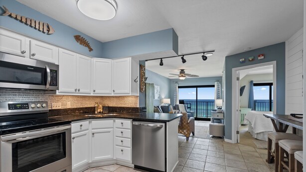 A modern kitchen with white cabinets, a stainless steel microwave, and an oven. There is a small island with a dishwasher. The room has a blue accent wall and a view of the ocean through a large window. - Sundunes 133 in Navarre Beach | Beachfront | Pool | My Beach Getaways (Navarre)