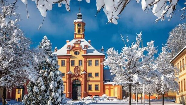 Ein großes, historisches Gebäude mit einem markanten Turm und einer symmetrischen Fassade, umgeben von schneebedeckten Bäumen und blauem Himmel.