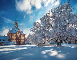 Paisaje nevado con un edificio grande que presenta una torre destacada y un árbol en primer plano.