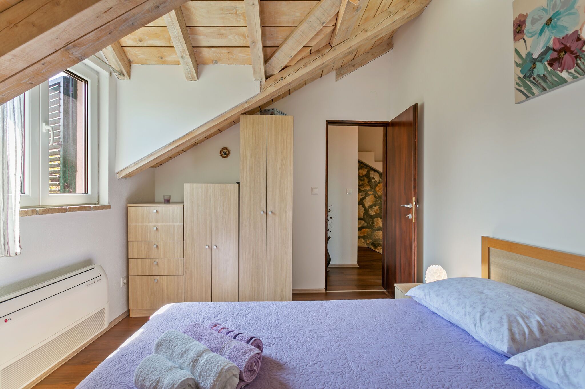 A bedroom with a sloped ceiling, a bed with a white and purple bedspread, a wooden wardrobe, and a window with blinds.