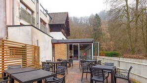 Eine Terrasse im Freien mit Tischen und Stühlen, eine Steinmauer und Blick auf Bäume und Hügel.