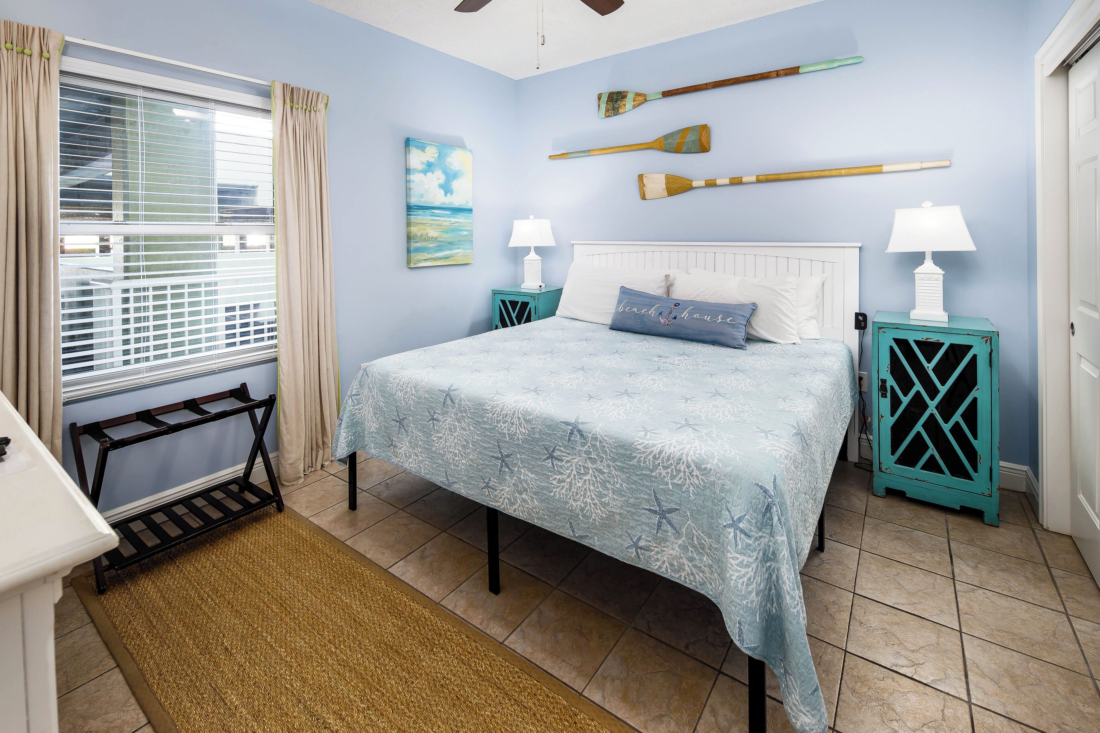 A bedroom with a bed, two nightstands, a window with curtains, a ceiling fan, and a wall-mounted paddleboard.
