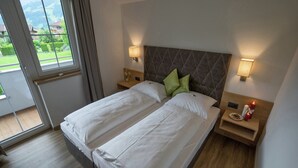 A double bed with white linens, two bedside tables with lamps, a wooden headboard, and a view of greenery through a window.