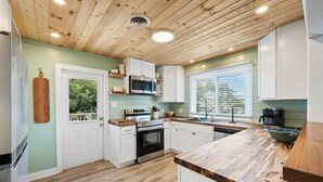 A modern kitchen with wooden ceiling, white cabinets, a built-in oven, and a window with blinds. - Spacious Second-Row Home Across from Beach Access (Georgetown)