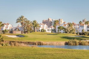 A golf course with a water hazard, palm trees, and residential buildings in the background.