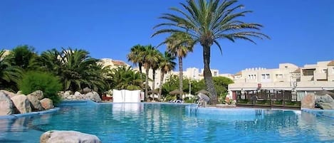 A clear blue pool with palm trees and rocks, surrounded by buildings.