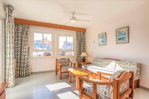 Ein Zimmer mit einer Couch, zwei Sesseln, einem kleinen Tisch, einem Deckenventilator und einem Fenster mit Vorhängen.