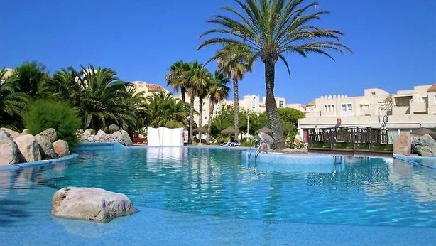 A pool area with clear blue water, surrounded by rocks and palm trees, with buildings in the background.