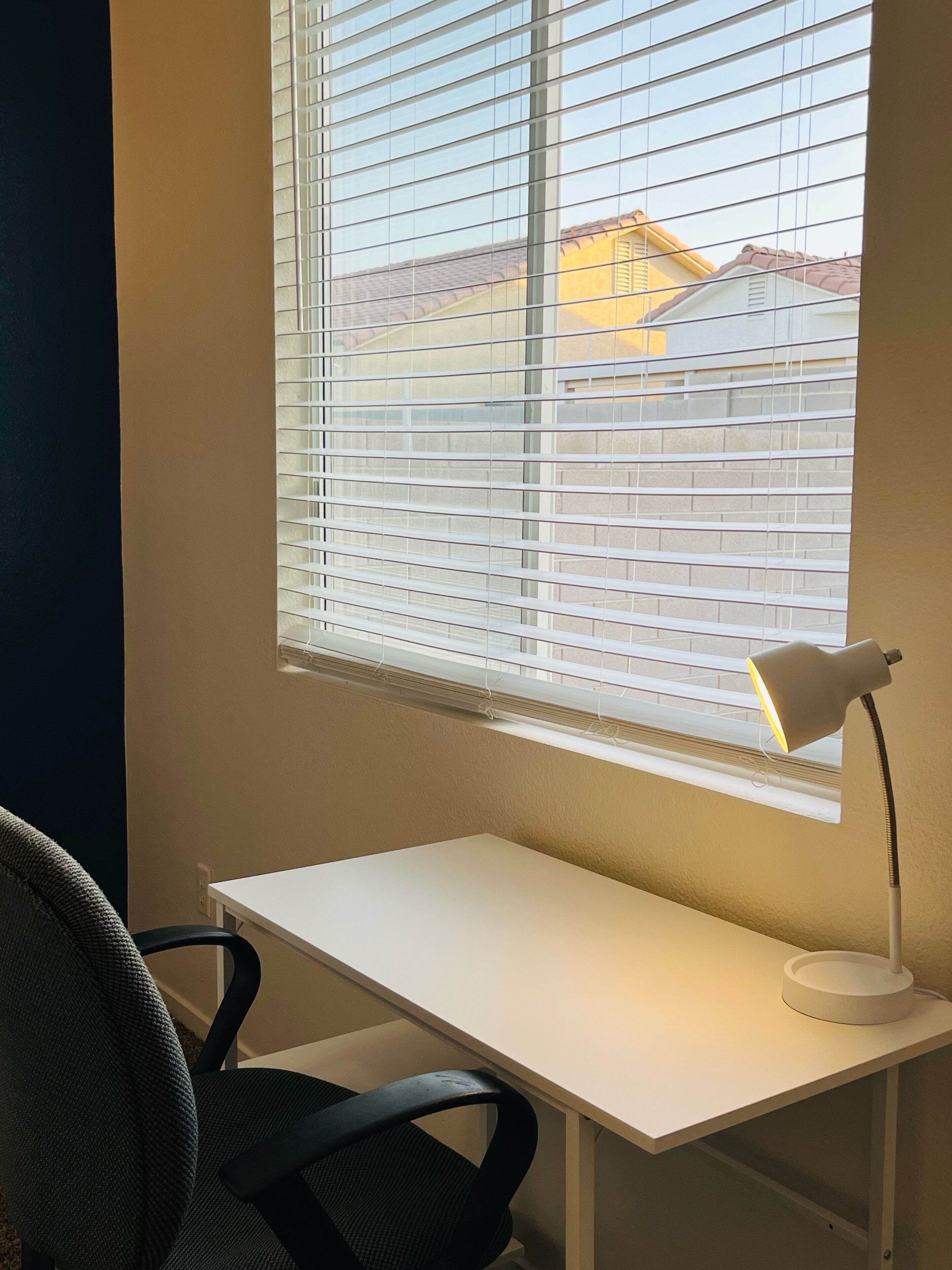 Una habitación con un escritorio blanco, una silla de oficina negra, una ventana con persianas y una lámpara de escritorio.
