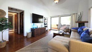 A spacious living room with a large flat-screen TV, a beige sofa, and a coffee table. There is a wooden cabinet with decorative items and a large window with curtains.