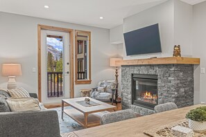 A living room with a fireplace, a television mounted on the wall, and a door leading to a balcony. - Super comfy in Spring Creek ~ Canmore 208TML (Canmore)