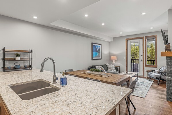 A modern kitchen with a large island, a built-in sink, and a dining area with a wooden table and chairs. - Super comfy in Spring Creek ~ Canmore 208TML (Canmore)