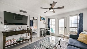A living room with a TV, a ceiling fan, a glass coffee table, a sofa, and a dining area with chairs and a table. - Beach Hair; Don't Care - Waves, Views, and Zero Worries! (Galveston)