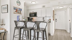 A kitchen with white cabinets, a microwave, and two bar stools. - Beach Hair; Don't Care - Waves, Views, and Zero Worries! (Galveston)