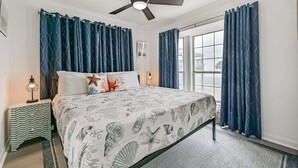 A bedroom with a bed, curtains, a ceiling fan, a nightstand, and a window. - Beach Hair; Don't Care - Waves, Views, and Zero Worries! (Galveston)