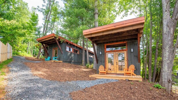 A rustic cabin with a wooden exterior, two porches with Adirondack chairs, and a gravel driveway.
