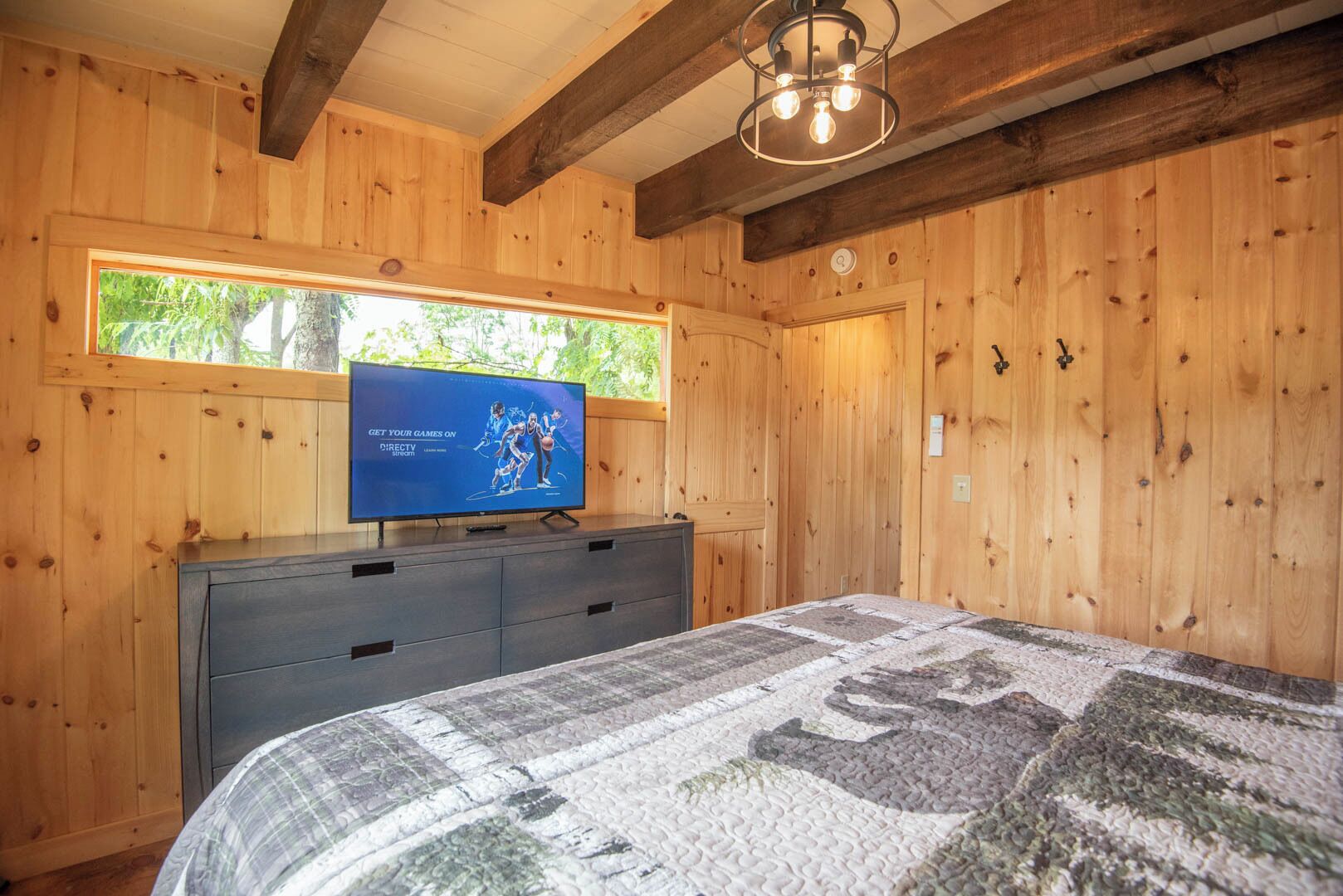 A wooden cabin bedroom with a bed, a TV on a dresser, and a window with a view of trees.