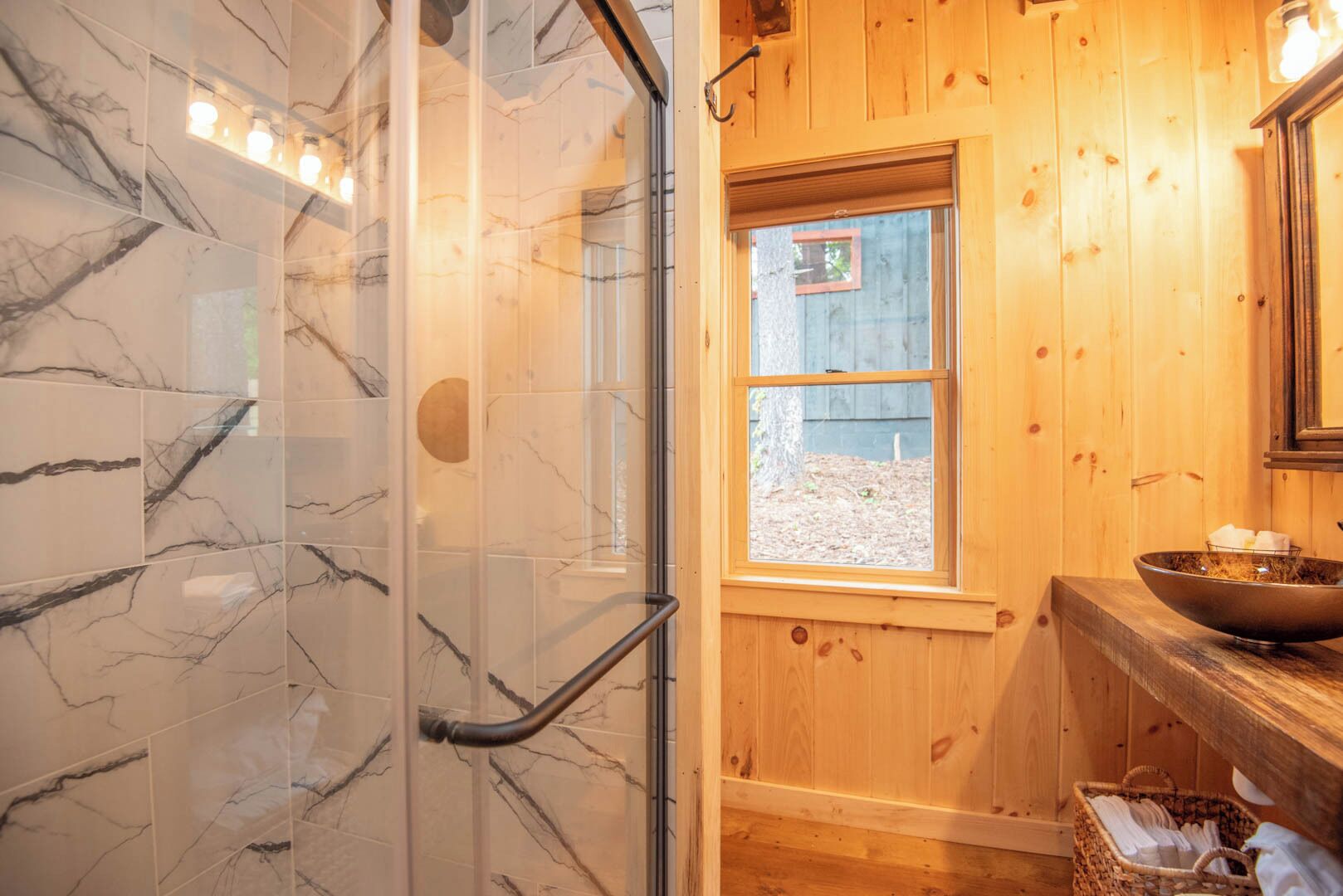 Ein Badezimmer mit Marmorfliesen, einer Glasdusche, einem Holzbadezimmermöbel und einem Fenster mit Blick ins Freie.