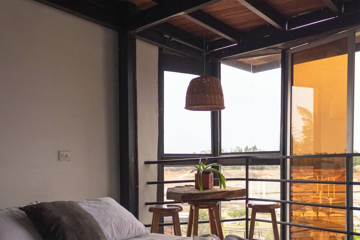Un dormitorio con una cama grande, una mesita con una planta y vistas a través de un ventanal amplio.