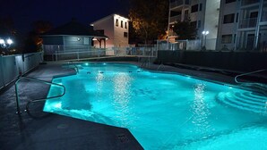 A swimming pool at night with clear water, surrounded by a fence and a building in the background.