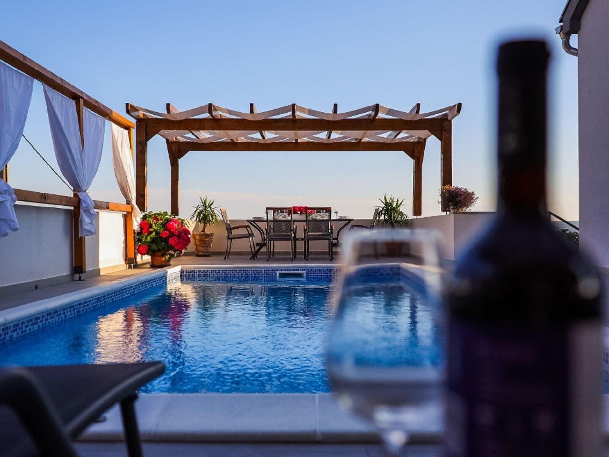 A pool area with a pergola, seating, and a bottle in the foreground.