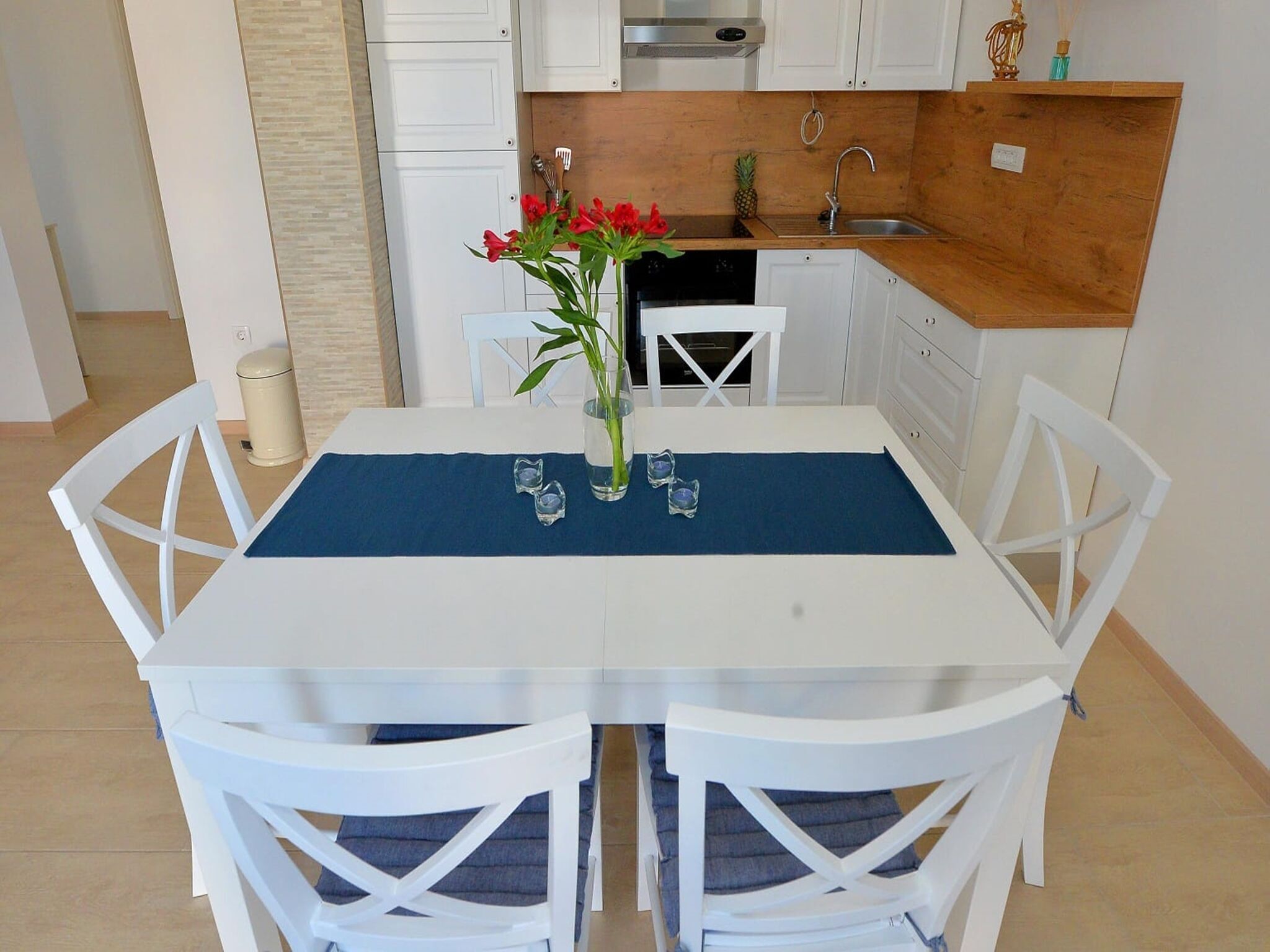 Een eethoek met een tafel voor twee personen, witte stoelen, een vaas met bloemen en een keuken op de achtergrond.