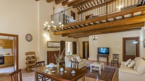 A spacious living area with a wooden dining table, chairs, a sofa, a TV, and a staircase leading to an upper level.