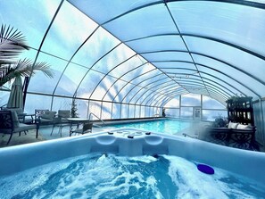 Een groot overdekt zwembad met glazen dak, omringd door ligbedden en een whirlpool.