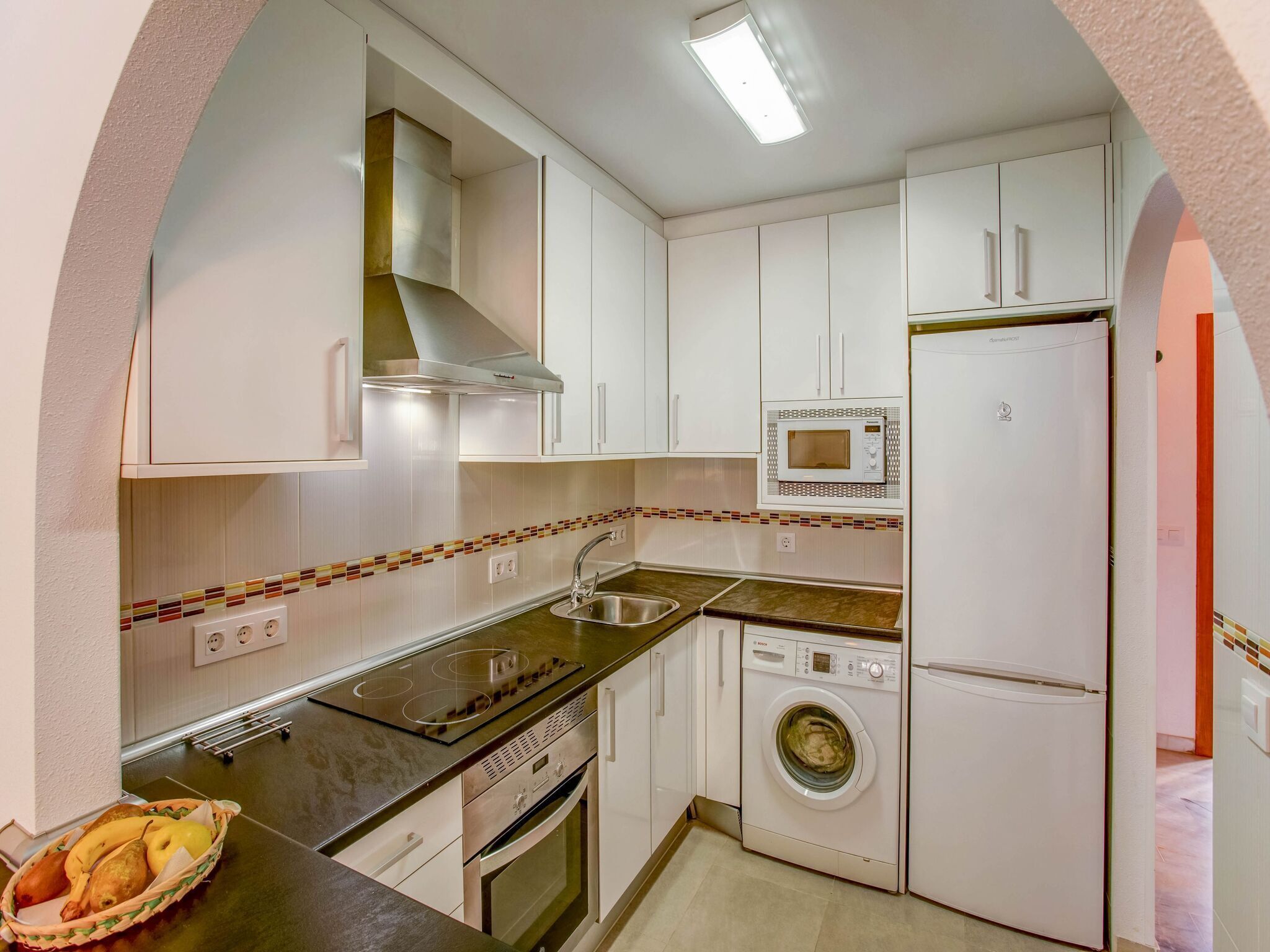 Cocina compacta con armarios blancos, microondas integrado, lavadora y estufa.